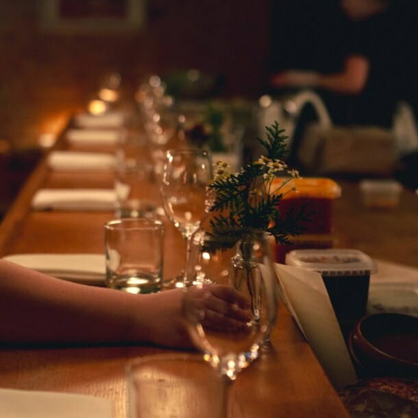Gedeckter Holztresen bei schummrigen Licht, auf den eine Frauenhand eine kleine Blumenvase mit Kamille stellt.