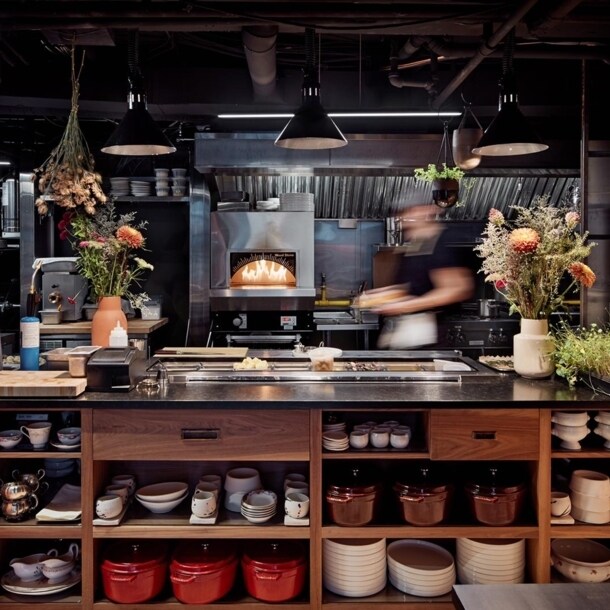 Blick in die Küche, wo im Ofen ein Feuer brennt und neben der unverputzten Backsteinwand eine Anrichte mit Blumensträußen und frischen Kräutern steht.