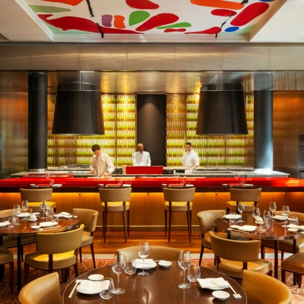 Interieur eines elegant-modernen Restaurants mir runden gedeckten Holztischen und offenem Tresen, in dem drei Köche stehen.