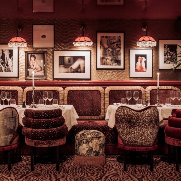 Gedeckte Tische in einem in dunklen Farben gehaltenem Interieur mit Sesseln und Teppich mit Animal-Muster sowie Fotografien an der Wand und Fransenlampen an der Decke.