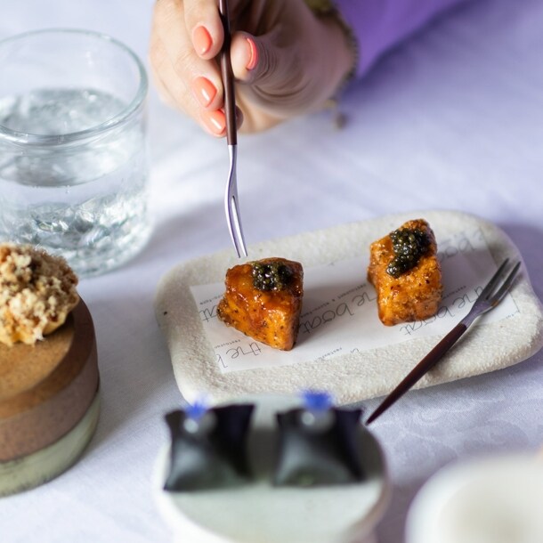 Eine Hand führt eine Gabel zu einem kunstvoll angerichteten Dessert auf einem weiß eingedeckten Restauranttisch.