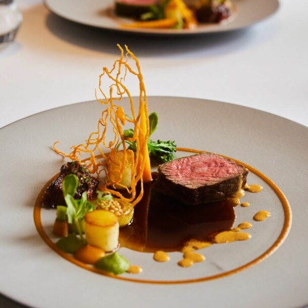 Kunstvoll arrangiertes Gericht mit einem Stück Fleisch auf einem großen weißen Teller auf einem weißen Tisch.