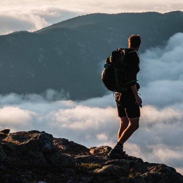 Wanderer mit Rucksack steht auf felsigem Gipfel und blickt auf nebelverhangene Berge bei Sonnenuntergang