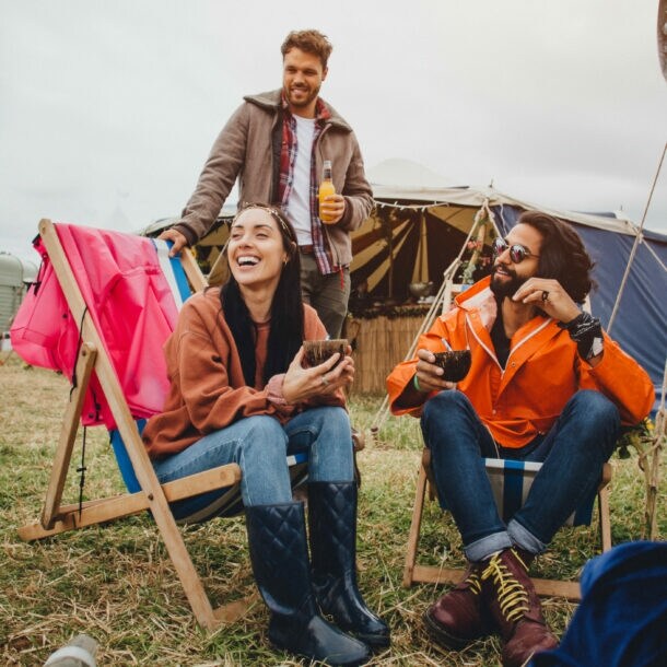 Drei Personen sitzen und stehen entspannt auf einem Festivalgelände mit Zelten im Hintergrund, lachen und trinken Getränke.