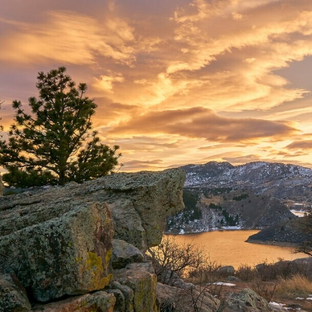 Felsige Landschaft mit Nadelbäumen und See bei Sonnenuntergang unter bewölktem Himmel