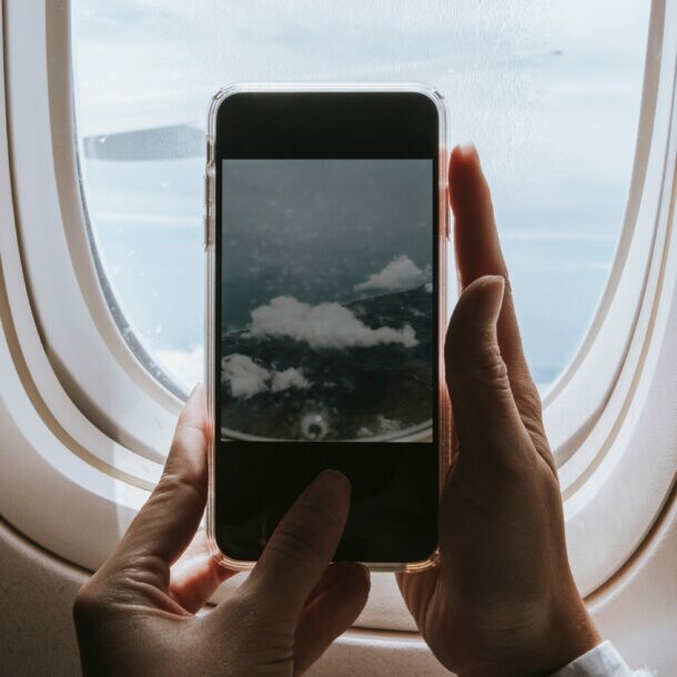 Hände halten ein Smartphone vor einem Flugzeugfenster, das den Himmel mit Wolken zeigt.