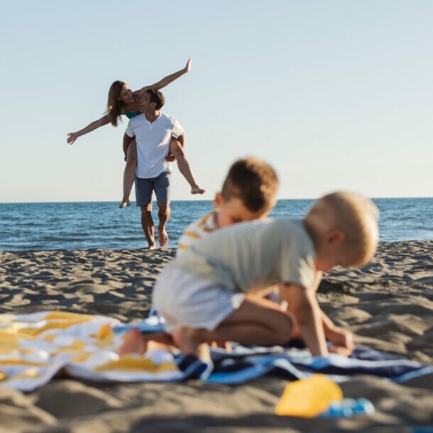 Zwei spielende Kinder auf einem Strandtuch am Strand, im Hintergrund trägt ein Mann eine Frau auf dem Rücken am Meer
