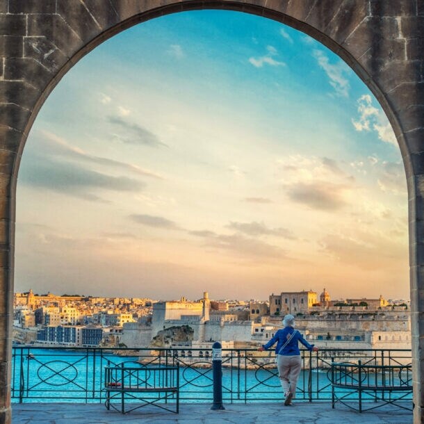 Blick durch einen großen Steinbogen auf eine Stadt am Wasser bei Sonnenuntergang, eine Person lehnt am Geländer