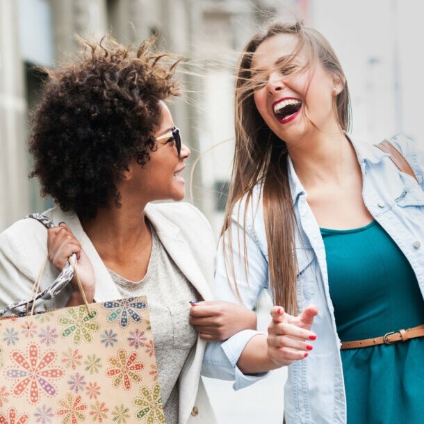 Zwei lachende Frauen mit Einkaufstüten auf dem Gehweg einer Straße.
