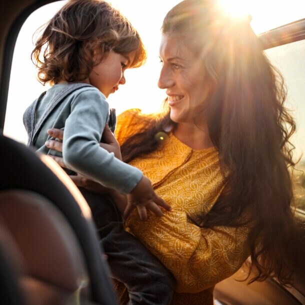 Frau in gelbem Kleid hält lächelnd ein kleines Kind im Auto bei Sonnenuntergang