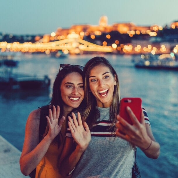 Zwei junge Frauen machen abends am Flussufer ein Selfie, im Hintergrund beleuchtete Brücke und Stadtlichter
