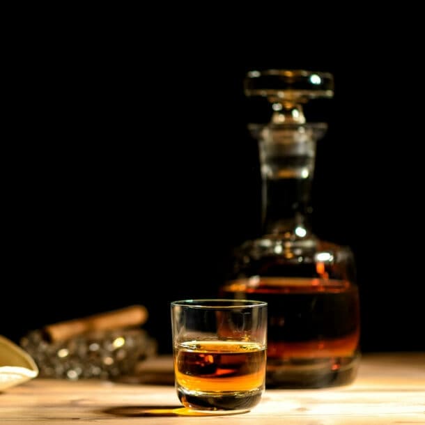 Beiger Strohhut mit schwarzem Band neben einem Glas und einer Karaffe mit braunem Getränk auf einem Holztisch vor schwarzem Hintergrund