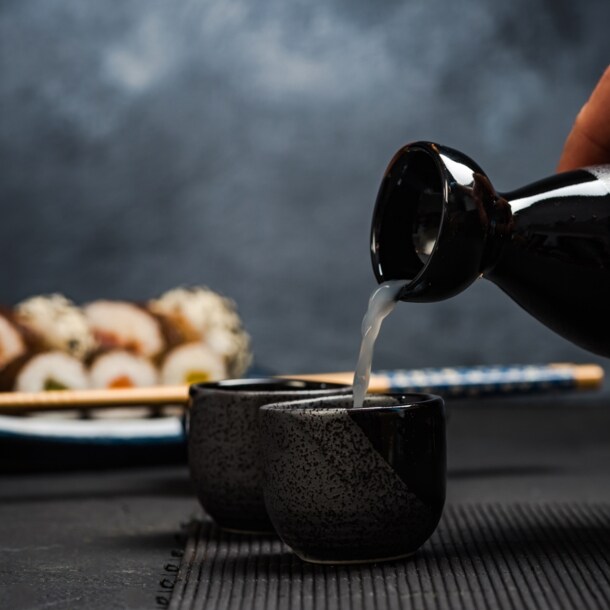 Hand gießt Sake aus einer schwarzen Karaffe in eine kleine schwarze Tasse, im Hintergrund Sushi und Essstäbchen