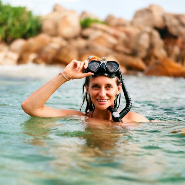 Frau mit Schnorchelmaske im Wasser vor felsiger Küste, hebt die Maske lächelnd an