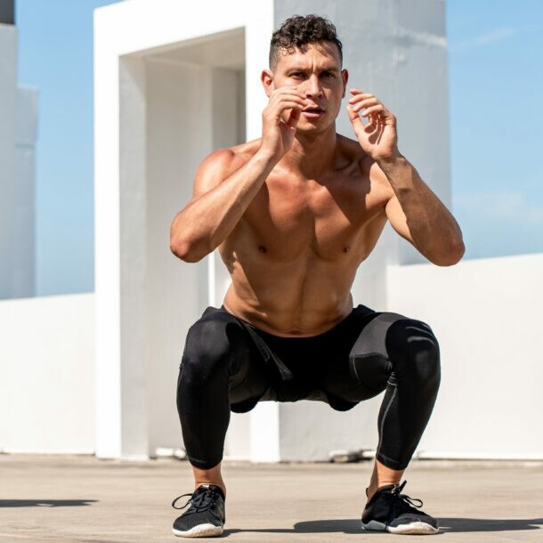Drei Männer machen Kniebeugen im Freien vor weißer Architektur, zwei sind oben ohne, einer trägt ein schwarzes Shirt und eine Kappe