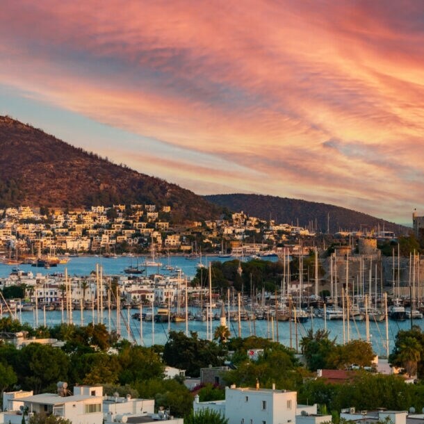 Sonnenuntergang über einer Küstenstadt mit Hafen, vielen Segelbooten und Bergen im Hintergrund.