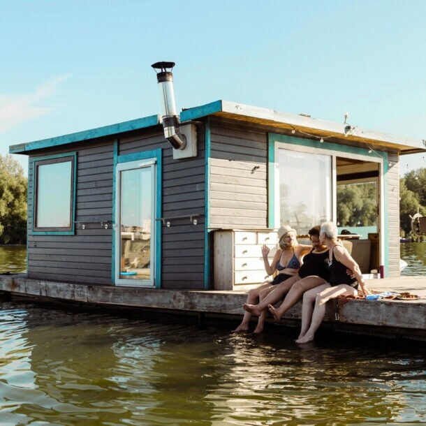 Drei Frauen in Badekleidung sitzen am Rand eines Pontons mit Hausboot und lassen ihre Füße ins Wasser baumeln bei Sonnenschein.