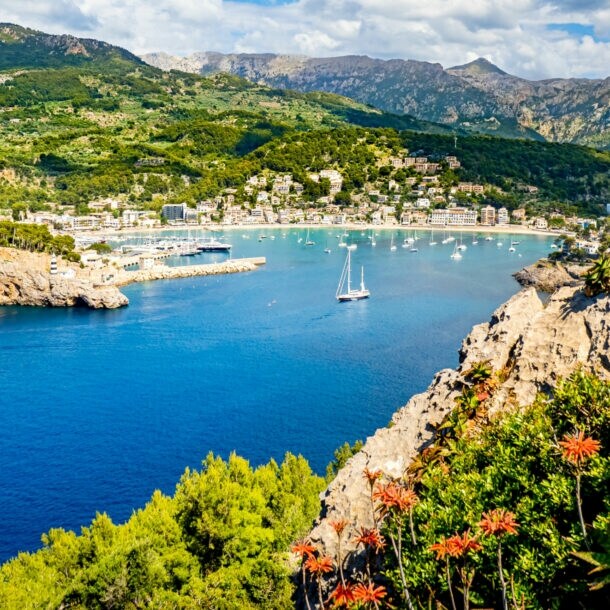 Bucht mit klarem blauem Wasser, Segelboot und Yachten, umgeben von grünen Hügeln und Bergen unter bewölktem Himmel