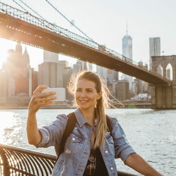 Frau macht Selfie vor der Brooklyn Bridge und der Skyline von New York bei Sonnenuntergang