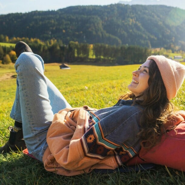 Eine Frau ruht sich auf einer Wiese in der Sonne aus, dabei liegt sie auf ihrem Wanderrucksack.