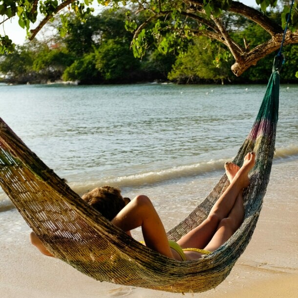 Person entspannt in einer Hängematte am Strand unter einem Baum mit Blick auf das Meer