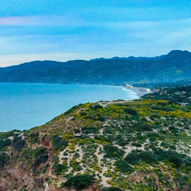 Klippen mit grüner Vegetation an der Küste bei Sonnenuntergang, dahinter Berge und ein Dorf.