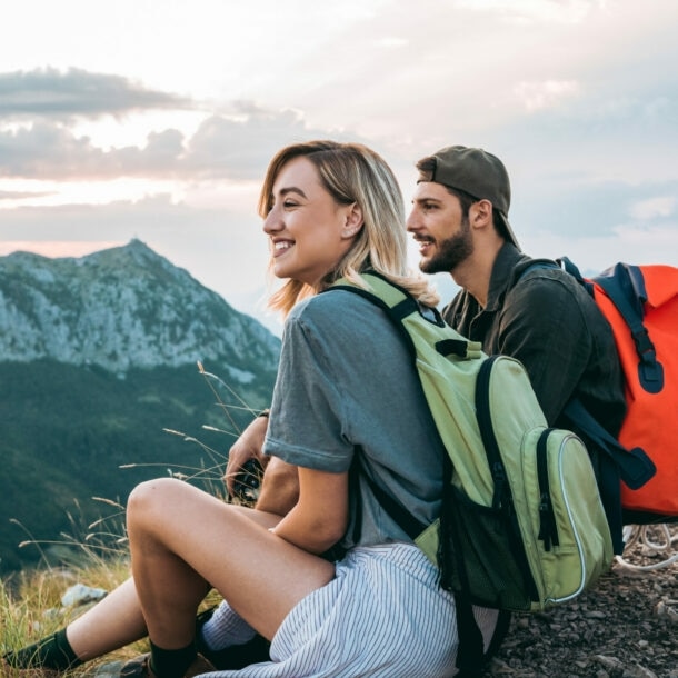 Ein Pärchen sitzt mit Rucksäcken auf den Rücken in der Natur und schaut auf umliegende Berge