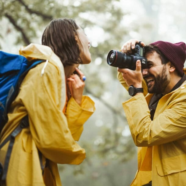 Mann mit roter Mütze und gelbem Regenmantel fotografiert lachende Frau mit blauem Rucksack und gelbem Regenmantel im Wald