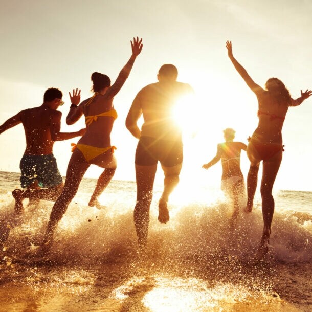 Gruppe von sechs jungen Menschen läuft im Sonnenuntergang ins Meer, Wasser spritzt um sie herum.