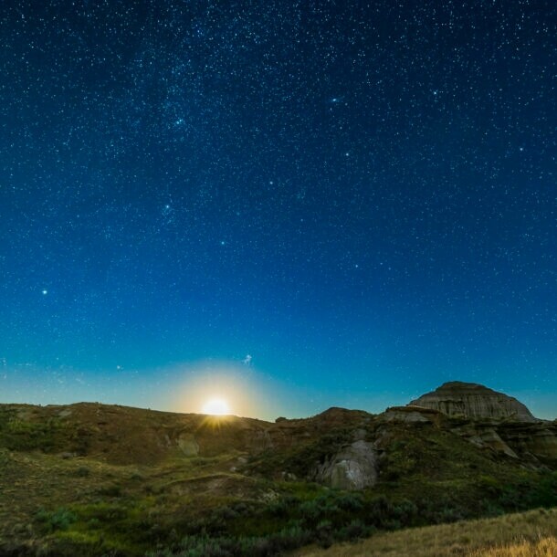 Klarer, tiefblauer Sternenhimmel mit aufgehendem Mond über einer felsigen Graslandschaft.