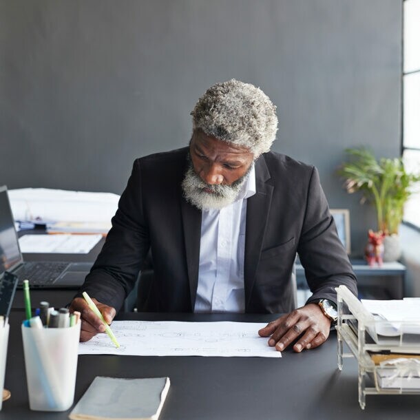 Ein älterer Mann arbeitet fokussiert im Büro an einer technischen Zeichnung.