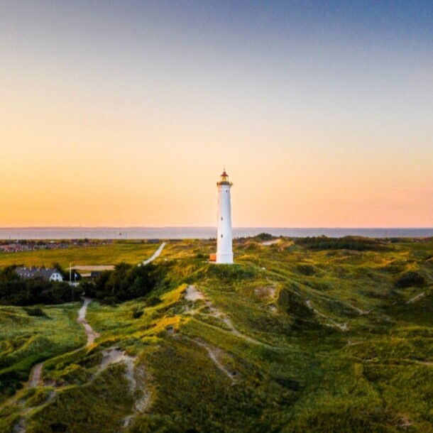 Weißer Leuchtturm auf grünen Dünen bei Sonnenuntergang mit Dorf und Meer im Hintergrund.