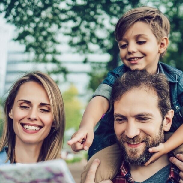 Vater trägt Sohn auf Schultern, Mutter hält Karte, sie stehen in einem Park mit Bäumen und Gebäuden im Hintergrund
