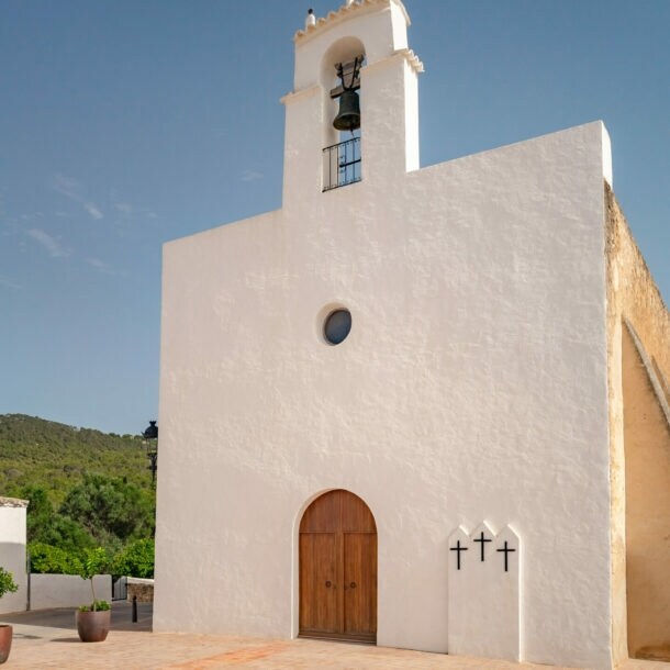 Weiße Kirche mit Glockenturm und drei Kreuzen an der Wand, umgeben von Gebäuden und Bergen unter blauem Himmel