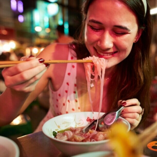 Nahaufnahme einer jungen, lächelnden Frau beim Essen eines asiatischen Nudelgerichtes mit Essstäbchen auf einem Straßenmarkt bei Nacht.