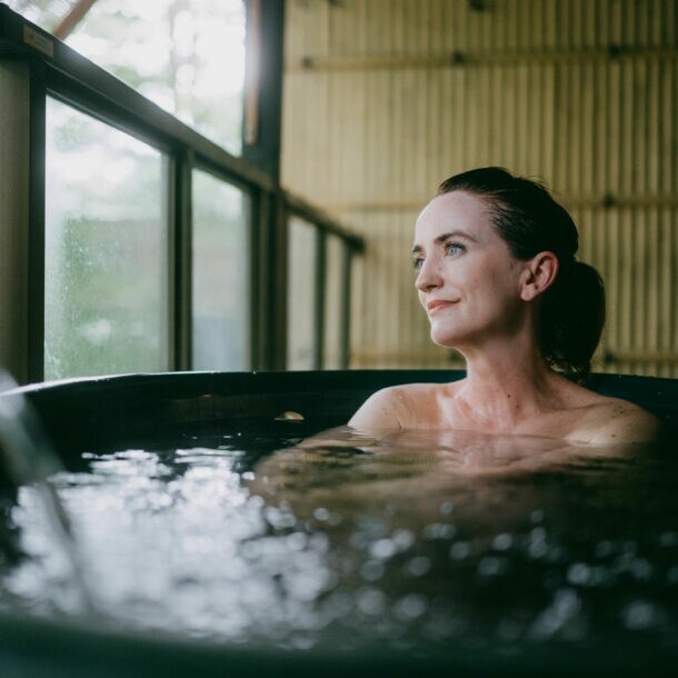 Eine Frau sitzt in einem Thermalbad aus dunklem Stein in einem japanischen Spa mit Bambuswand und schaut zufrieden durch ein Fenster hinaus.