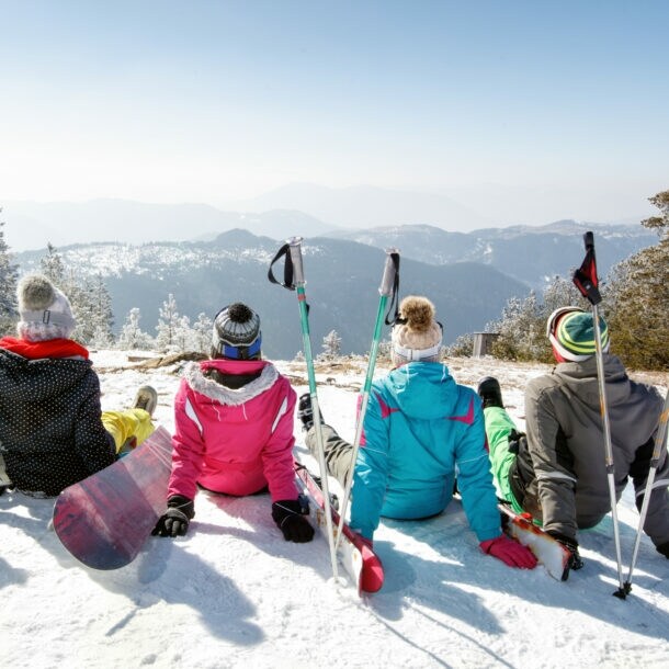 Fünf Personen mit Snowboards und Ski sitzen mit Blick auf eine weitere Berglandschaft auf einem Berg im Schnee.