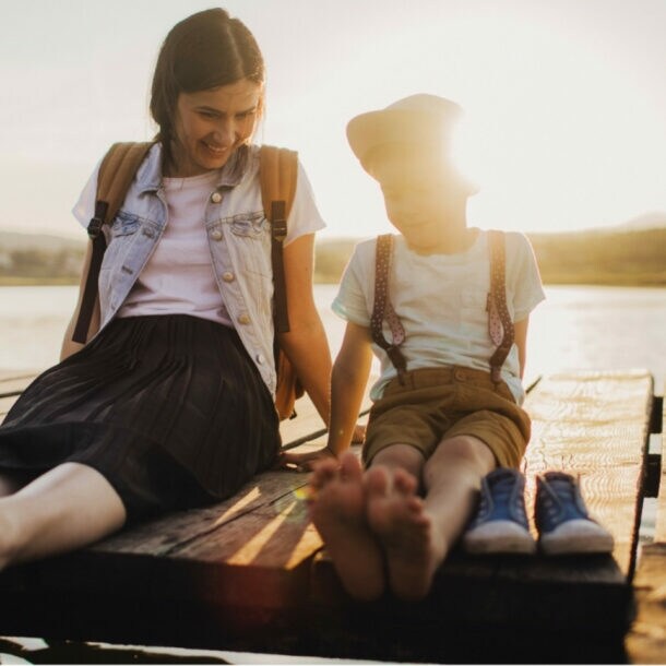Mutter und Kind sitzen barfuß auf einem Holzsteg am See, die Sonne scheint im Hintergrund