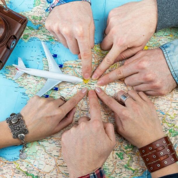 Sechs Personen, von denen nur die Hände zu sehen sind, zeigen auf einen Punkt auf einer Landkarte. Daneben liegen ein alter Fotoapparat und ein Modell eines Flugzeugs.