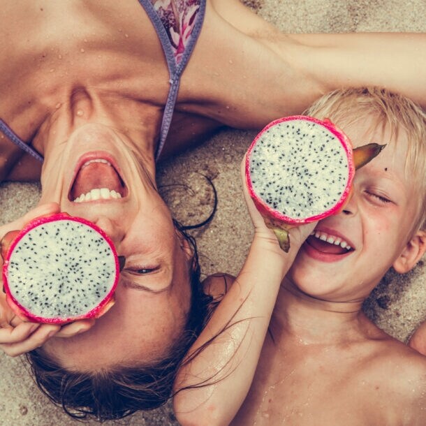Eine Frau liegt mit einem Kind im Sand, beide halten eine tropische Frucht vor einem ihrer Augen.