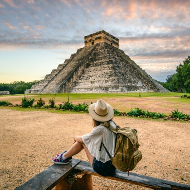 Person mit Hut und Rucksack sitzt auf einer Bank und blickt auf die Pyramide von Chichén Itzá bei Sonnenuntergang