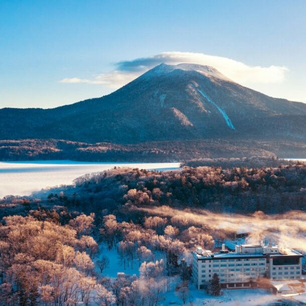 Verschneite Landschaft mit Wald und Gebäuden vor einem großen Berg unter klarem Himmel bei Sonnenaufgang