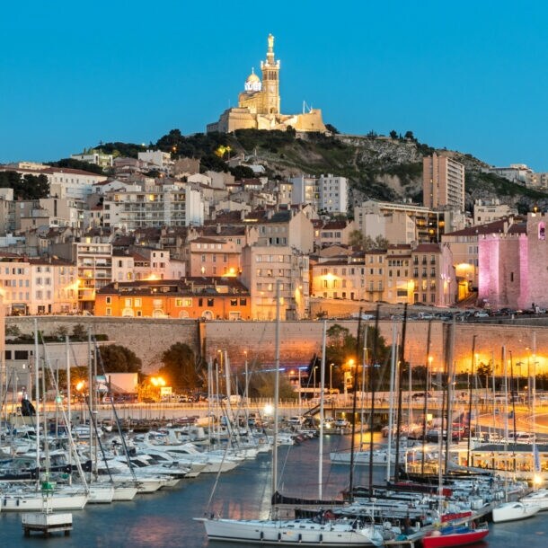 Abendliche Hafenansicht von Marseille mit Segelbooten im Wasser und der beleuchteten Kirche Notre-Dame de la Garde auf einem Hügel im Hintergrund