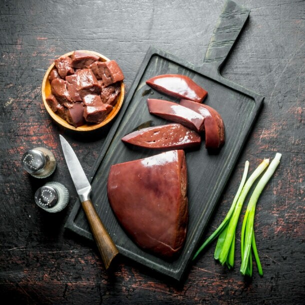 Rohe Leberstücke auf schwarzem Schneidebrett mit Messer, Frühlingszwiebeln und Gewürzstreuern auf dunklem Untergrund