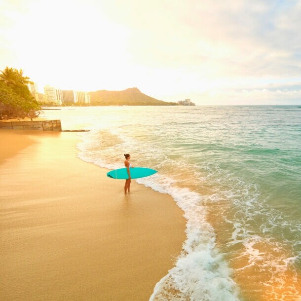 Eine Frau mit türkisfarbenem Surfboard unterm Arm steht am frühen Morgen am leeren Strand von Waikiki.