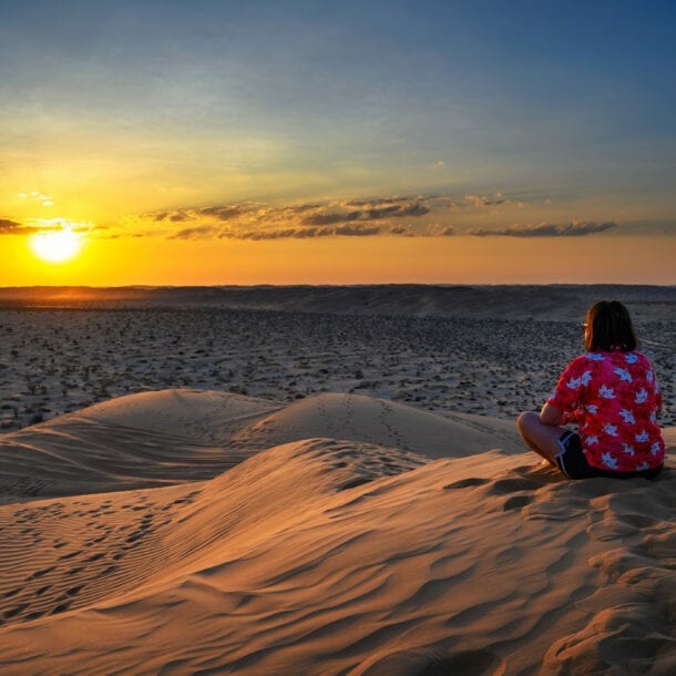 Person in rotem Hemd mit weißen Mustern sitzt auf Sanddüne und blickt auf Sonnenuntergang über Wüste