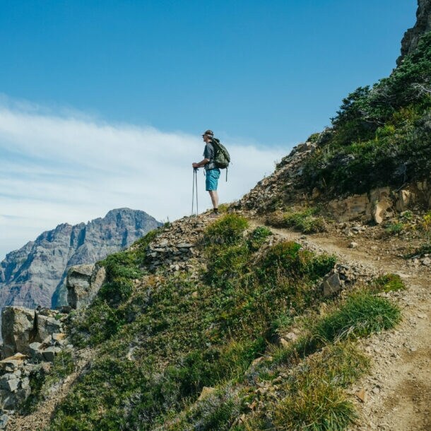 Wanderer mit Rucksack und Wanderstöcken steht auf einem Bergpfad und blickt auf eine Berglandschaft unter blauem Himmel