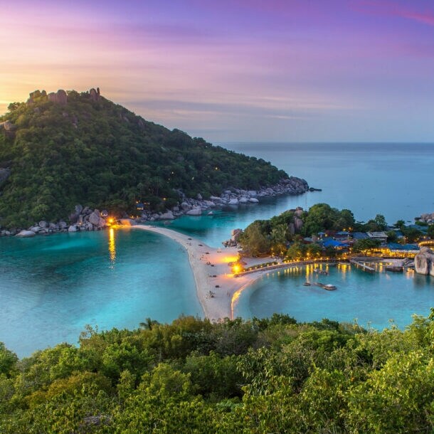 Blick auf eine tropische Insel mit bewaldetem Hügel, Sandbank und beleuchteten Gebäuden bei Sonnenuntergang