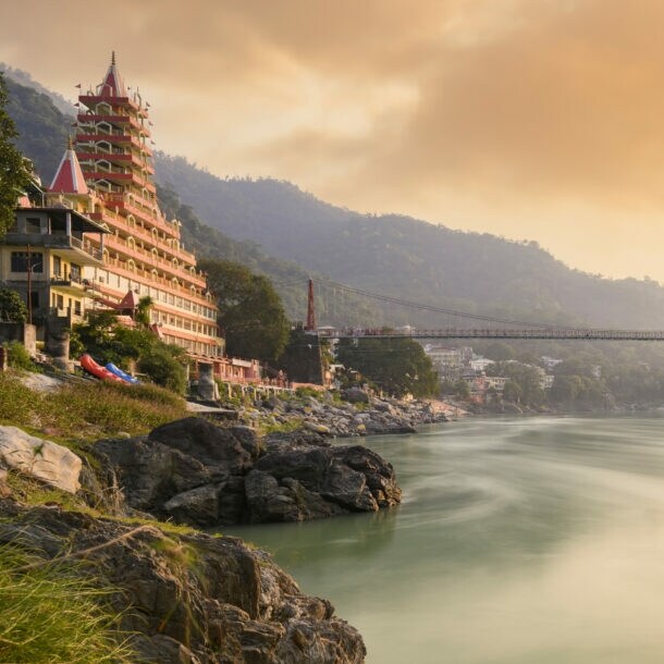 Fluss mit hängender Fußgängerbrücke und mehrstöckigem Tempelgebäude am Ufer bei Sonnenuntergang in bergiger Landschaft