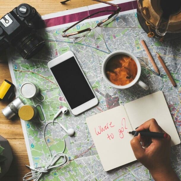 Hand schreibt 'Where to go?' in Notizbuch auf Landkarte mit Kamera, Smartphone, Kaffeetasse, Brille, Reisepass und Münzen